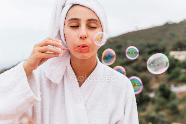Mujer joven en albornoz blanco y toalla que sopla burbujas de jabón durante la sesión de spa en el balcón - foto de stock