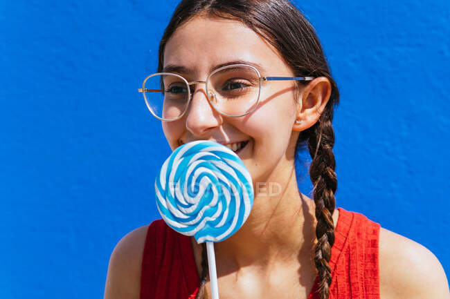 Позитивна чарівна жінка, що стоїть з солодким льодяником на вулиці в сонячний день на синьому фоні і дивиться в сторону — стокове фото
