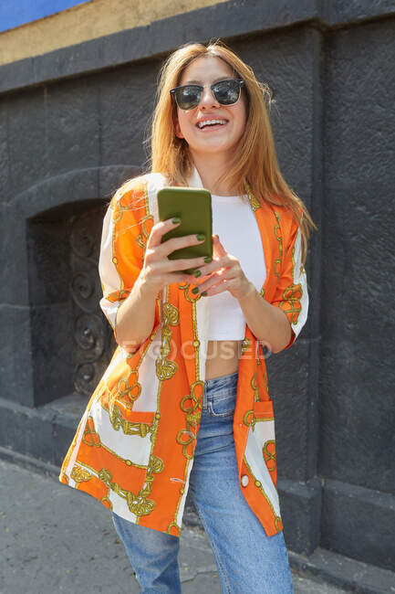 Trendy allegra donna in denim vestito in piedi in strada e la navigazione sui social media sul telefono cellulare — Foto stock