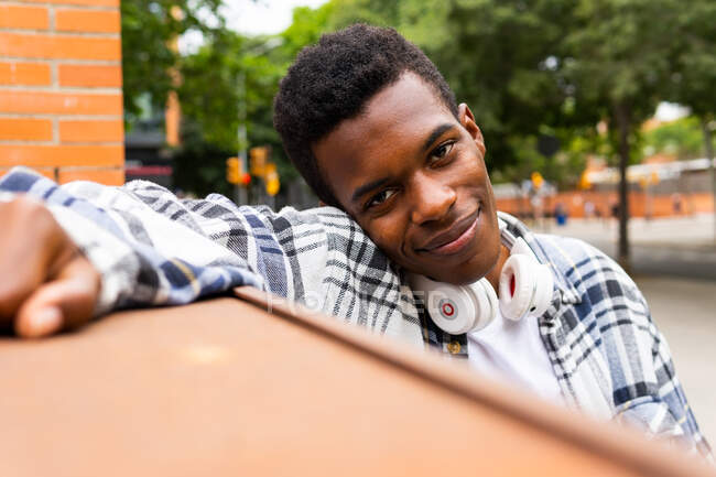 Alegre afroamericano hipster masculino en camisa a cuadros y con auriculares inalámbricos en el cuello de pie cerca de la construcción de la ciudad y mirando a la cámara - foto de stock