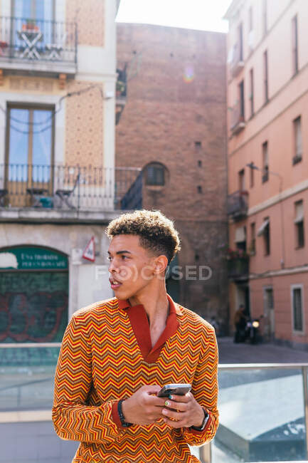 Joven hispano con cabello afro en elegante traje colorido navegando por el teléfono móvil mientras está parado en la barandilla cerca del edificio urbano a la luz del sol - foto de stock