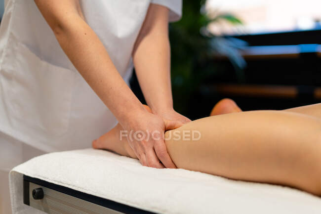 Massaggiatrice irriconoscibile ritagliata in vestaglia bianca massaggiando il vitello della paziente durante la sessione di fisioterapia in clinica — Foto stock