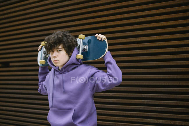 Неэмоциональный подросток в повседневной одежде смотрит в камеру, стоя со скейтбордом за головой на улице на размытом фоне. — стоковое фото