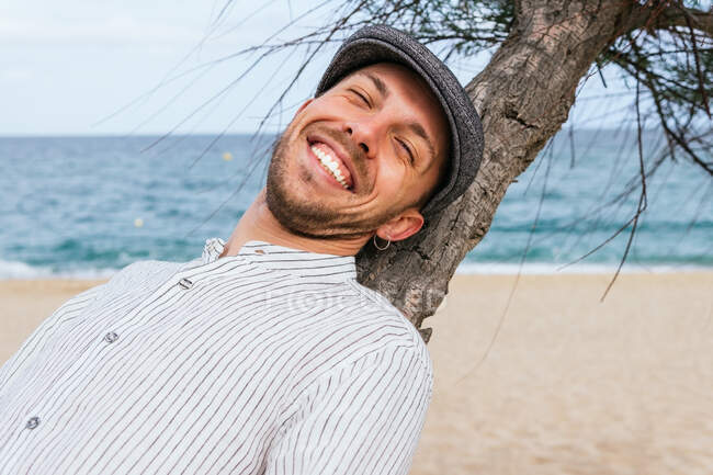 Glücklicher junger bärtiger Kerl in stylischem gestreiftem Hemd und Mütze lehnt sich auf trockenen Baumstamm zurück und genießt den Sommerurlaub am Sandstrand am Meer — Stockfoto