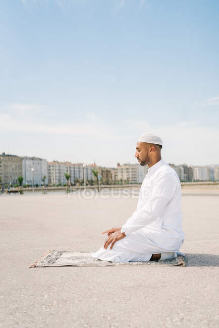 Ragazzo pieno maschio islamico in abiti bianchi tradizionali in ginocchio sul tappeto e pregando contro il cielo blu sulla spiaggia — Foto stock