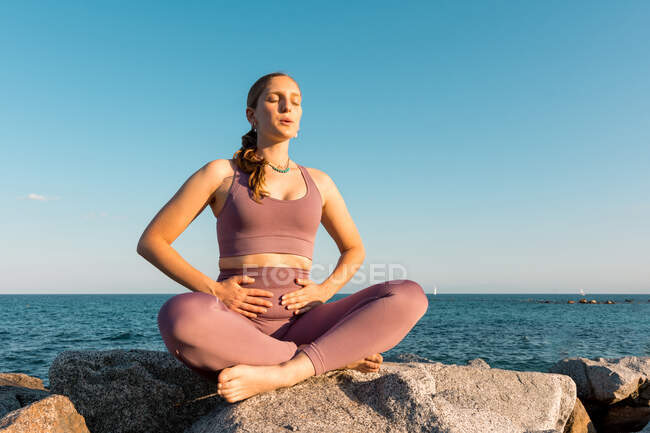 Mujer serena haciendo yoga en pose de loto mientras respira durante la meditación en la roca en la orilla del mar - foto de stock