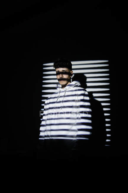 Jeune millénaire masculin méconnaissable en tenue décontractée debout dans une pièce sombre près d'un mur avec de l'ombre jalousie — Photo de stock