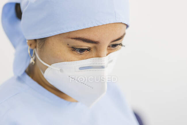 Cosecha joven médico femenino en máscara estéril y gorra mirando hacia abajo en el trabajo en el hospital - foto de stock