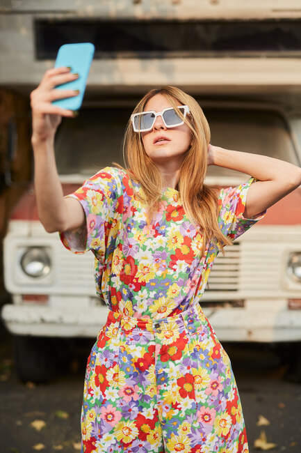 Female in trendy summer clothes and sunglasses standing in street and taking self shot on mobile phone — Stock Photo