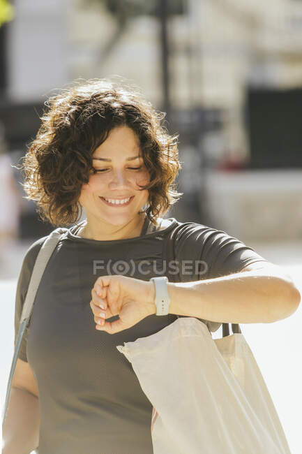 Happy young ethnic woman with brown curly hair in casual clothes checking time on wristwatch and smiling while waiting for meeting on city street in daytime — Stock Photo