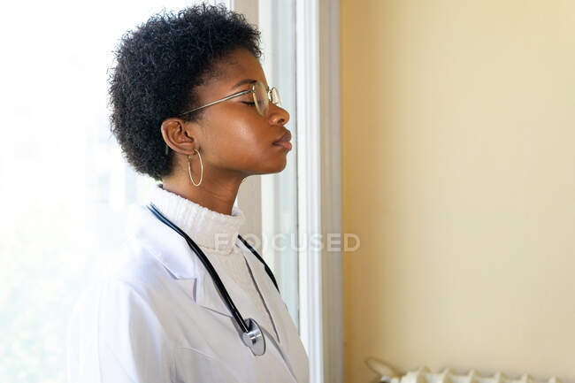 Вид сбоку компетентной молодой афроамериканской женщины-врача в белом медицинском халате и очках со стетоскопом закрытыми глазами во время пребывания в клинике — стоковое фото