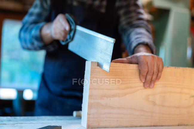 Crop unrecognizable male carpenter using sharp saw while cutting lumber plank at workbench in professional workshop — Stock Photo