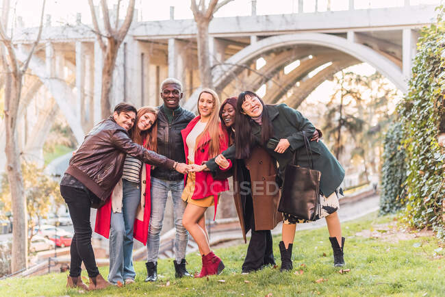 Компания счастливых разнообразных друзей обнимается, стоя на газоне в парке и глядя в камеру — стоковое фото