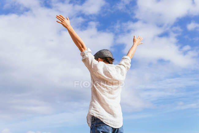 Low angle back view of unrecognizable carefree male traveler in stylish outfit raising arms and enjoying freedom against blue cloudy sky in summertime — Stock Photo