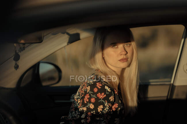 Retrato de uma bela jovem mulher loira pensativa sentada dentro de um carro olhando para a câmera ao pôr do sol — Fotografia de Stock