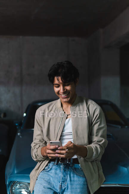 Jeune homme latino souriant en vêtements décontractés en utilisant le téléphone tout en s'appuyant sur une voiture vintage dans un parking — Photo de stock
