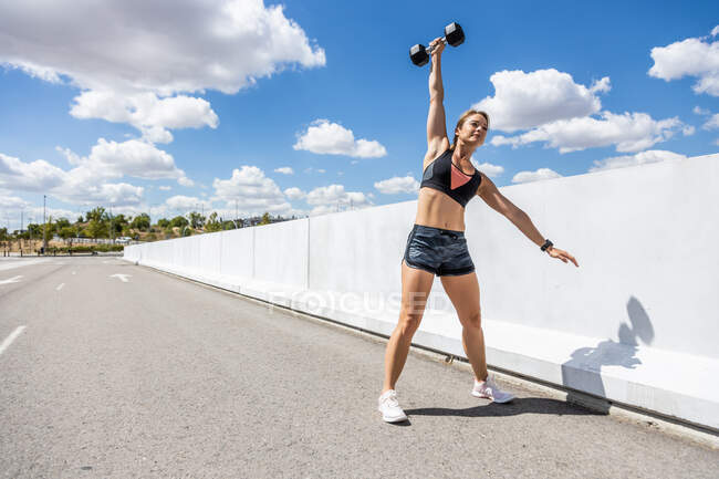 Молодая женщина тренируется с гантели на открытом воздухе, руки вверх, вид сбоку — стоковое фото