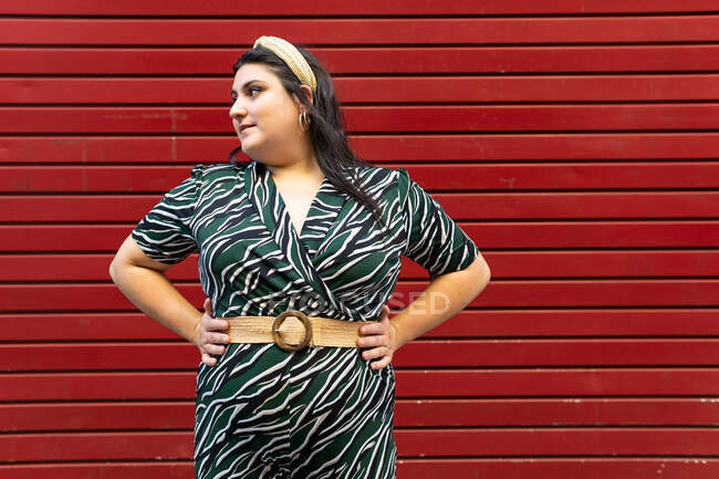 Ritratto di giovane bruna curvy allegra in elegante abito a righe e fascia mentre guarda lontano contro la parete rossa — Foto stock