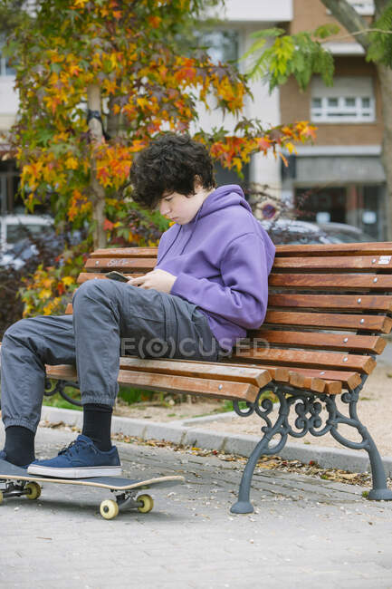 Вид сбоку сосредоточенного юноши-подростка, сидящего на деревянной скамейке с скейтбордом во время серфинга в интернете по мобильному телефону в парке на улице — стоковое фото