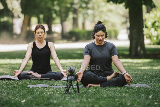 Mindful female bloggers with closed eyes sitting in Padmasana pose while recording video on photo camera in park — Stock Photo