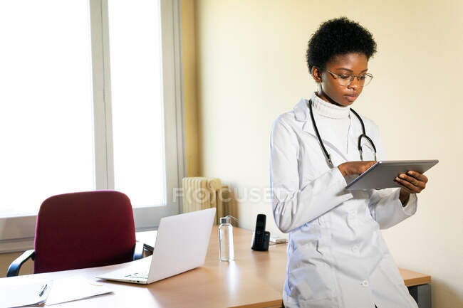 Jovem médica negra séria em casaco médico com estetoscópio trabalhando com tablet no escritório da clínica moderna — Fotografia de Stock