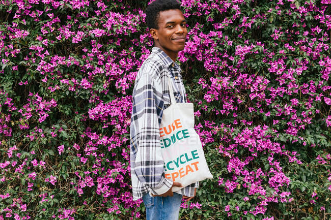Side view of cheerful African American male standing with eco friendly shopping bag in blooming summer garden and looking at camera — Stock Photo