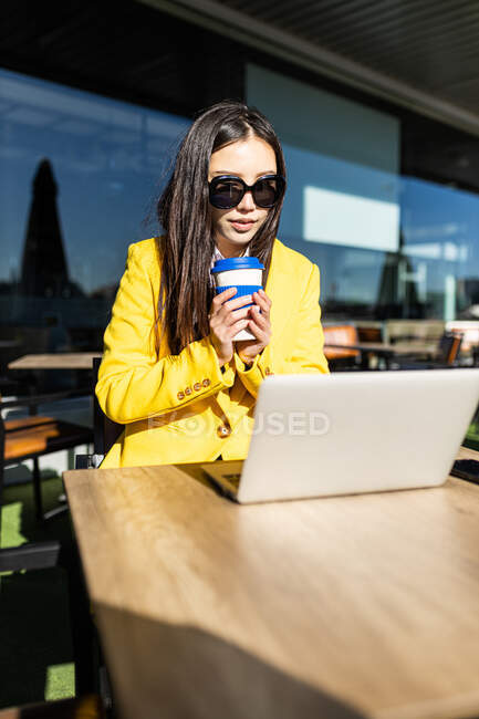 Азиатская деловая женщина в желтом пальто сидит за столом и пьет кофе со своим смартфоном и ноутбуком — стоковое фото