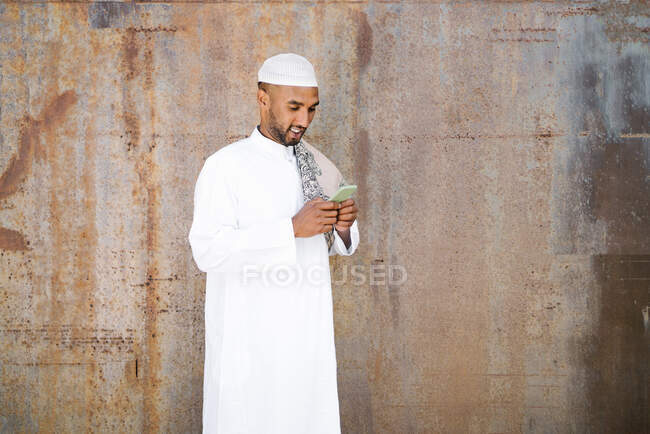 Cheerful Muslim male in traditional clothes smiling and browsing cellphone while standing near shabby wall on street — Stock Photo