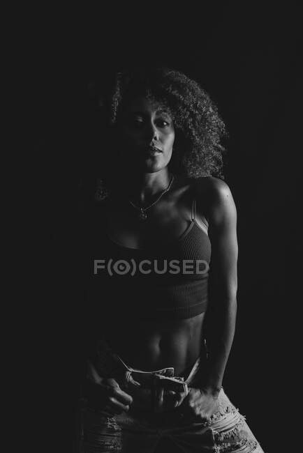 Curly haired African American female in trendy crop top and jeans standing with hand on waist on black background in studio — Stock Photo