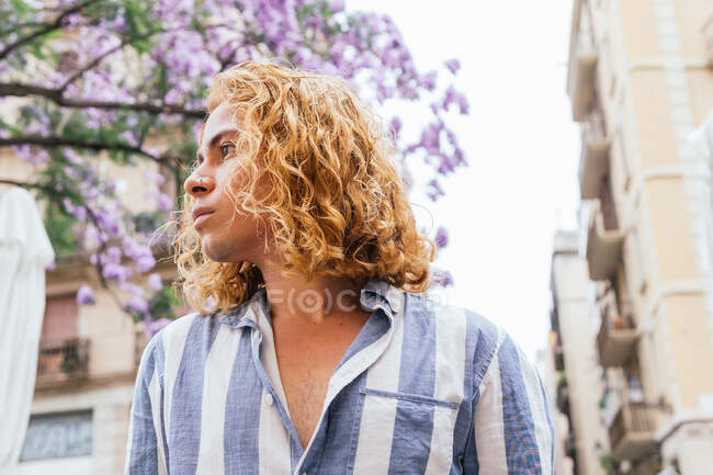 D'en bas de mâle curieux rêveur avec de longs cheveux ondulés debout dans la rue en été avec arbre à fleurs — Photo de stock