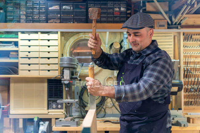 Side view of cheerful mature male master using hammer and chisel while creating wooden detail in carpentry workshop — Stock Photo