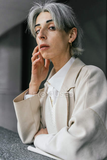 Серьезный трансгендер женщина в модном пальто опираясь на серый забор, стоя на улице глядя в камеру — стоковое фото
