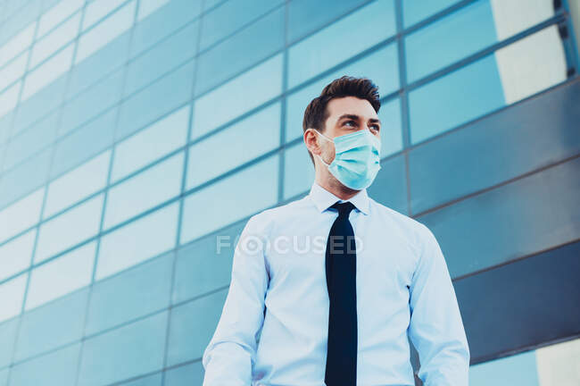 Знизу чоловічий підприємець у формальному носінні з медичною маскою, що дивиться далеко в місто під час пандемії коронавірусу — стокове фото