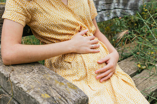 Crop anonymous pregnant female in summer dress touching tummy sitting on bench in the countryside in summer — Stock Photo