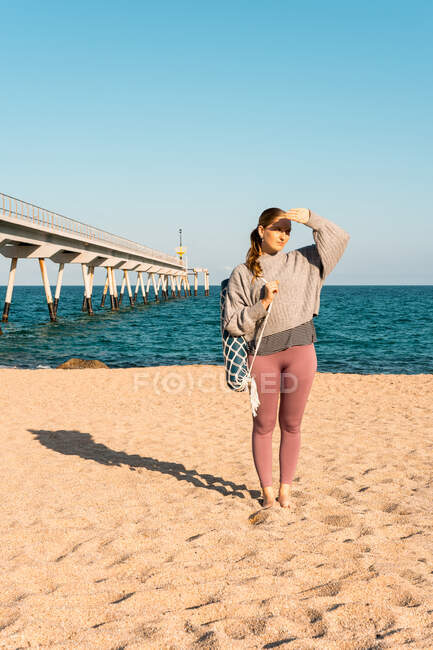 Cuerpo completo de joven hembra descalza en ropa deportiva con estera de yoga enrollada mirando a la distancia mientras está de pie en la playa de arena cerca del mar - foto de stock