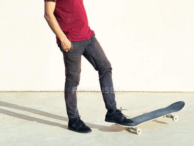 Обрезанный неузнаваемый мужчина-скейтбордист с руками в карманах, смотрящими на тротуар в солнечном свете на бежевом фоне — стоковое фото