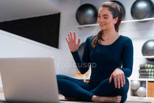 Ganzkörper positiver junger weiblicher Instruktor winkt mit der Hand, während er die Schüler während des Online-Yoga-Kurses per Videochat auf dem Laptop begrüßt — Stockfoto