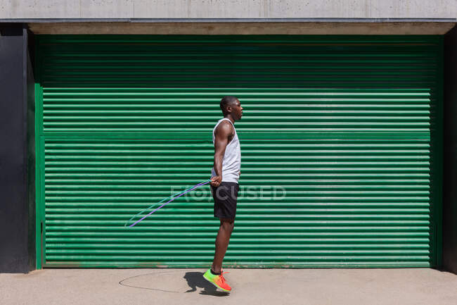 Atleta masculino afroamericano enfocado saltando la cuerda durante el entrenamiento cardiovascular en un día soleado en la ciudad - foto de stock