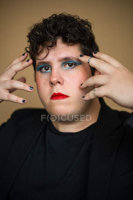 Emotionsloser androgyner Mann mit roten Lippen und hellem Lidschatten, der die Hände dicht vor dem Gesicht hält und in die Kamera blickt — Stockfoto
