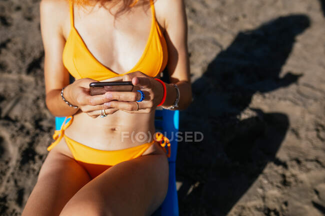 Anonimo giovane rossa donna utilizzando il telefono mentre seduto sulla sedia in spiaggia in una giornata di sole in estate — Foto stock