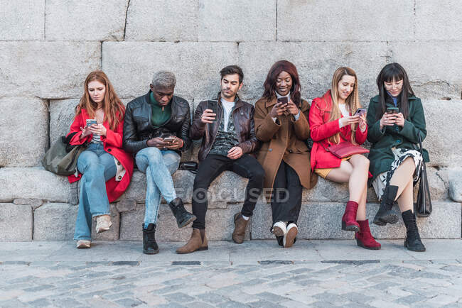 Compañía de diversos amigos sentados en la ciudad y utilizando teléfonos móviles mientras se enfría el fin de semana - foto de stock