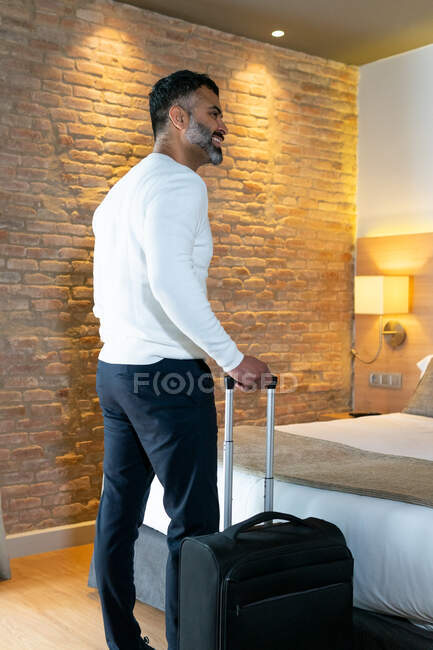 Visão traseira baixo ângulo de viajante masculino étnico positivo com bagagem em pé perto da cama no quarto do hotel — Fotografia de Stock