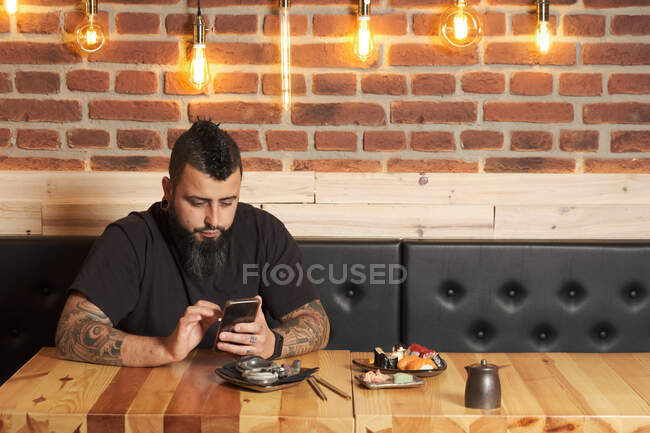 Masculino sentado à mesa com vários rolos de sushi e navegando no celular enquanto relaxa no restaurante japonês — Fotografia de Stock