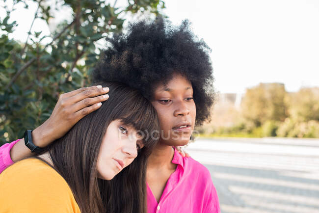 THigh angle of tranquil multiracial lesbian couple sitting on bench and embracing in city while looking away — Stock Photo