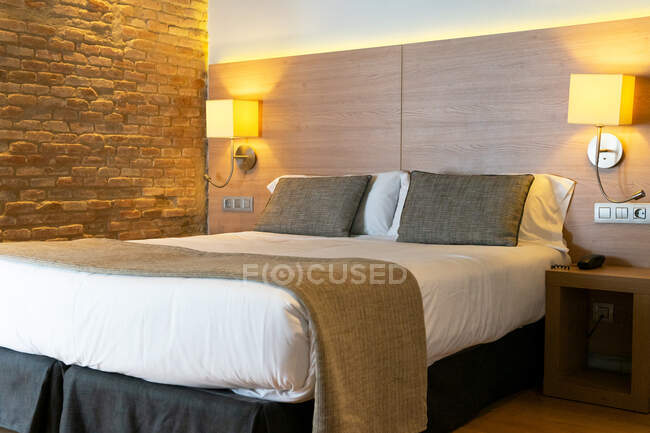 Comodo letto con cuscini posto su lenzuolo bianco con lampada appesa alla testiera in camera d'albergo — Foto stock