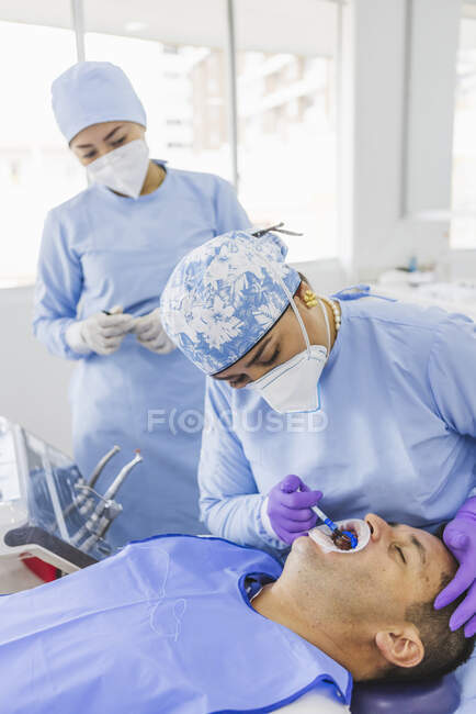 Doctora en guantes aplicando gel fluoruro en diente de hombre con retractor durante tratamiento dental en clínica - foto de stock