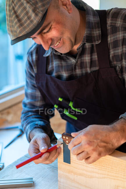 Trabajador de madera profesional positivo con lápiz y herramienta de medición que hace marcas en el tablero de madera mientras crea objetos de artesanía en el taller de carpintería - foto de stock