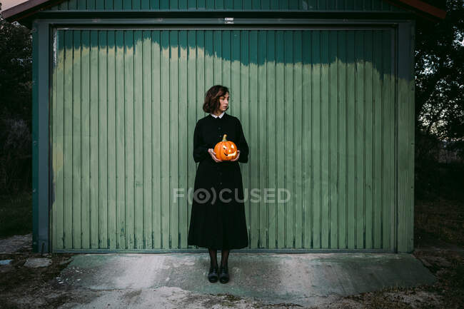 Mujer en vestido negro y con linterna de calabaza de Halloween de pie contra la pared de madera de la casa y mirando hacia otro lado - foto de stock
