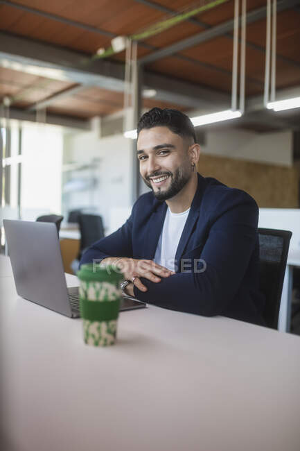 Веселий чоловік-підприємець дивиться на камеру під час роботи на робочому місці, сидячи за столом з ноутбуком — стокове фото