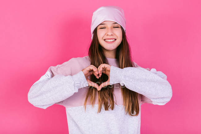 Усміхнена жінка-підліток із закритими очима та коричневим волоссям з головним убором для концепції ракових захворювань, що демонструє серце з руками на рожевому фоні — стокове фото